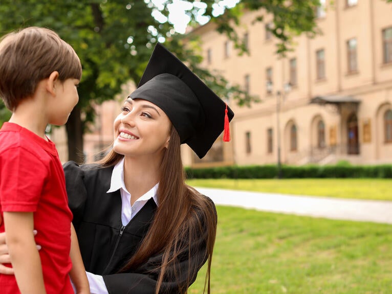 A smiling woman in a graduation cap and gown kneels next to a boy outside of a building.