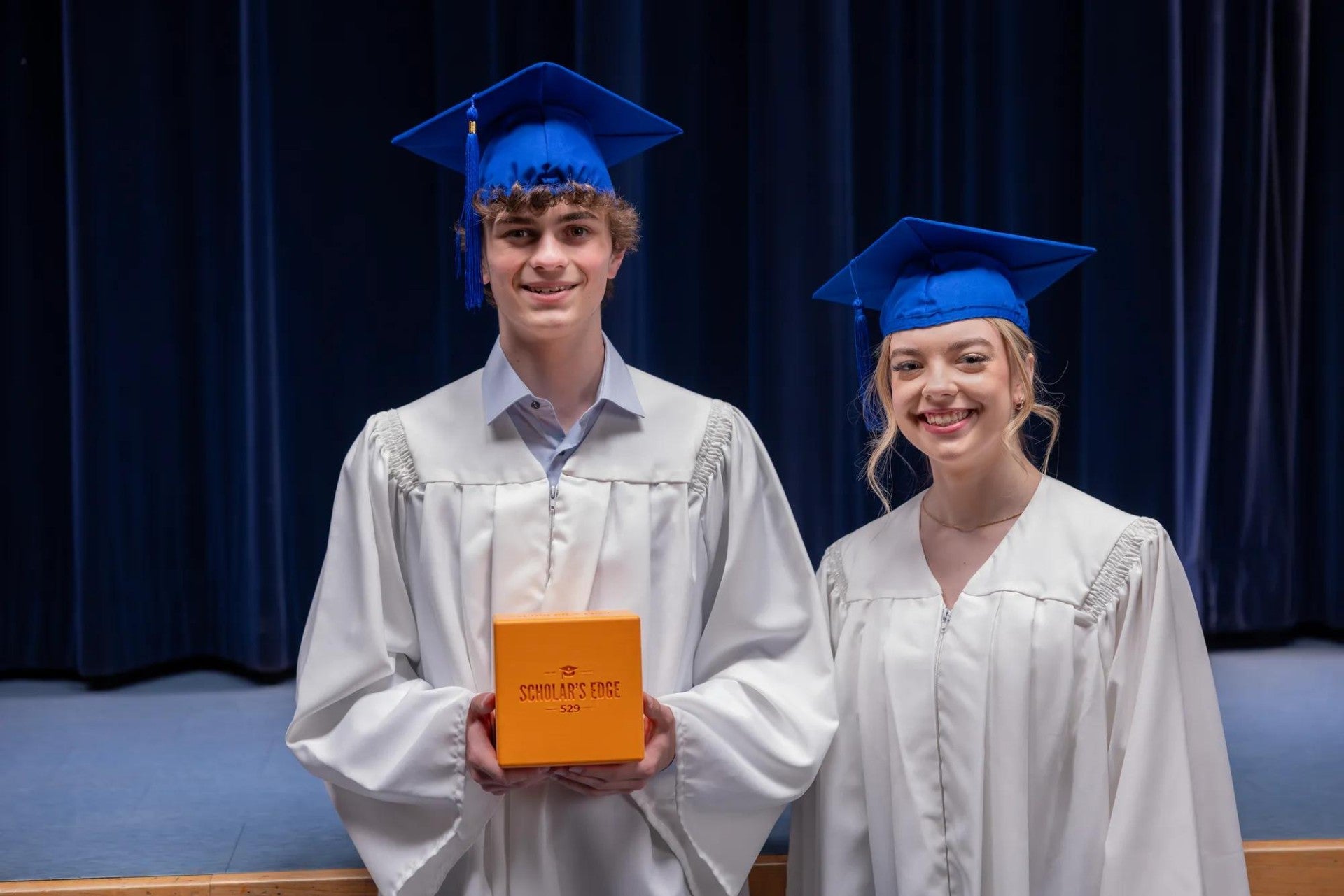Two teenagers in graduation caps and gowns holding the Scholar's Edge cube.